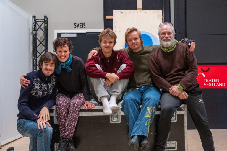 gruppebilde, fem glade tryner sit på ein flight case og held rundt kvarandre